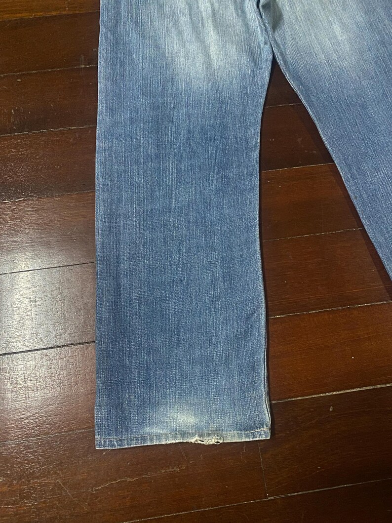 May include: Faded blue denim jeans with a straight leg cut. The jeans show wear and tear at the hem, with a slightly distressed appearance. The denim has a classic, washed look.