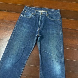 May include: A pair of classic blue denim jeans. The jeans have a button fly, five-pocket design, and a straight leg cut. The denim has a faded wash, giving the jeans a worn-in look. The jeans are laid flat on a wooden surface.