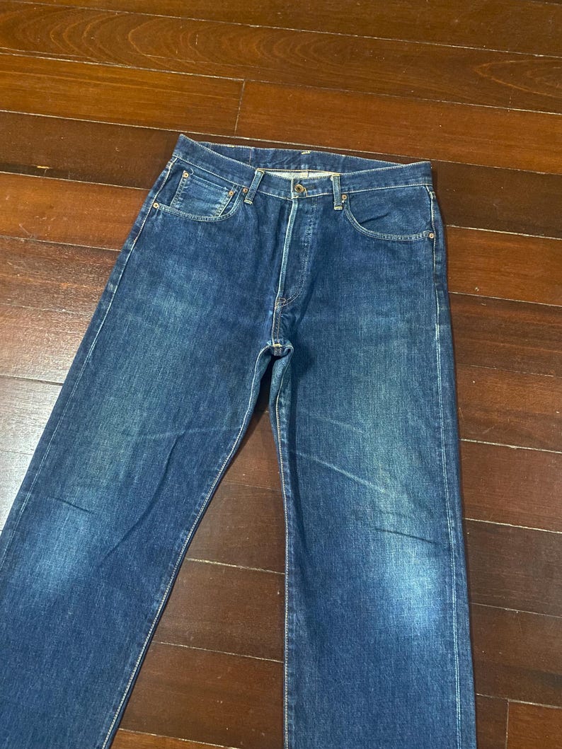 May include: A pair of classic blue denim jeans. The jeans have a button fly, five-pocket design, and a straight leg cut. The denim has a faded wash, giving the jeans a worn-in look. The jeans are laid flat on a wooden surface.
