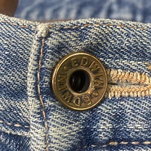 May include: Close-up of a brass button on blue denim fabric. The button is embossed with the word "EDWIN" in a circular pattern. The denim has visible stitching and a worn appearance.