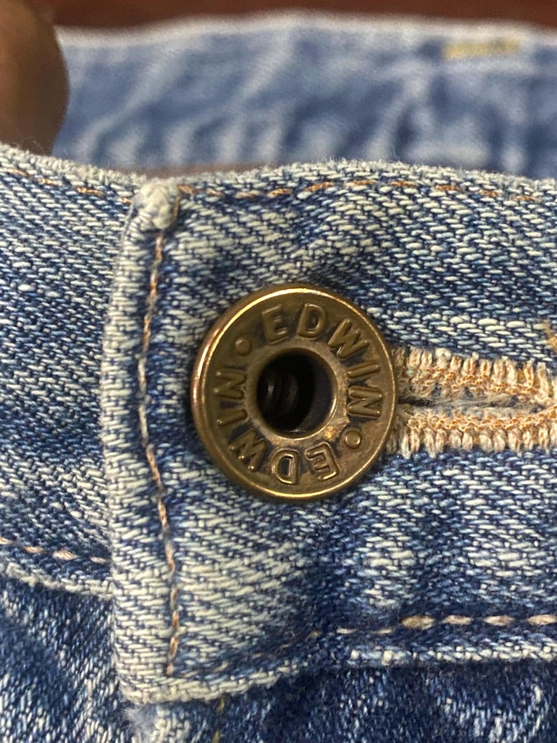 May include: Close-up of a brass button on blue denim fabric. The button is embossed with the word "EDWIN" in a circular pattern. The denim has visible stitching and a worn appearance.