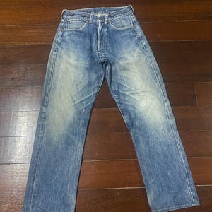 Może przedstawiać: Spodnie dżinsowe z wyblakłego niebieskiego denimu z rozporkiem na guziki, szlufkami na pasek i klasycznym pięciokieszeniowym designem. Dżinsy wykazują ślady zużycia i wyblaknięcia, nadając im wygląd vintage. Denim jest średniej gramatury.