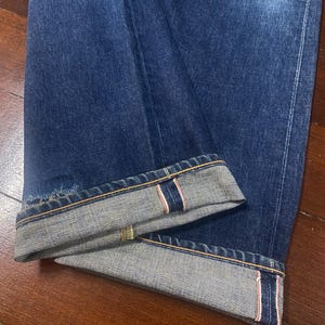 May include: Close-up of dark blue denim jeans with the cuffs rolled up. The rolled cuffs reveal a selvedge edge with red and white stitching. The jeans are folded on a wooden surface.
