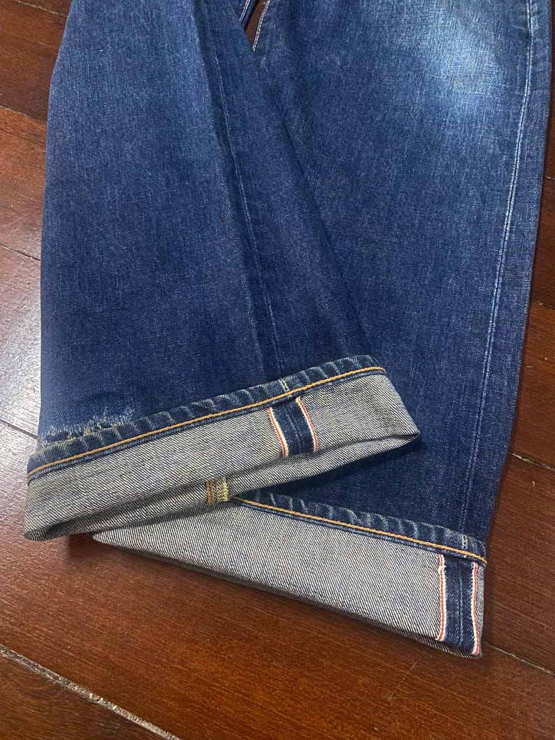 May include: Close-up of dark blue denim jeans with the cuffs rolled up. The rolled cuffs reveal a selvedge edge with red and white stitching. The jeans are folded on a wooden surface.