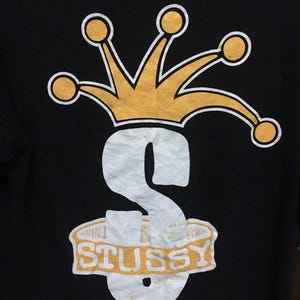 Puede incluir: Camiseta negra con un gráfico blanco y amarillo de un gorro de bufón con una corona encima de una 'S' estilizada con la palabra 'STUSSY' escrita en una pancarta debajo.