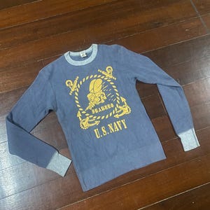 May include: Blue long-sleeve shirt with a waffle knit texture. The shirt features a yellow graphic of a sailor and the text "SEABEES" and "U.S. NAVY". The cuffs and collar are gray.