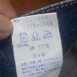May include: Close-up of a white tag with text and symbols, likely a clothing label. The tag reads "NO. ED-223 F535-0608 QUAL. 綿 100%" and includes washing instructions. The tag is attached to blue denim fabric.