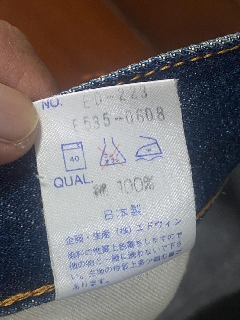 May include: Close-up of a white tag with text and symbols, likely a clothing label. The tag reads "NO. ED-223 F535-0608 QUAL. 綿 100%" and includes washing instructions. The tag is attached to blue denim fabric.