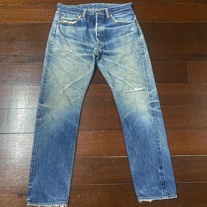 May include: Faded blue denim jeans with a button fly. The jeans have a worn appearance with distressed areas and light fading throughout. The straight-leg style features classic five-pocket design and belt loops.