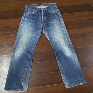 May include: Blue denim jeans with a button fly and faded areas on the thighs and knees. The jeans have a classic five-pocket design and a straight leg cut. The denim has a worn, vintage look.