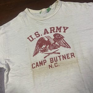 Pode incluir: Camiseta vintage creme com gola redonda. A frente apresenta um gráfico bordô de uma águia careca, as palavras "U.S. ARMY" e "CAMP BUTNER N.C." em um estilo desgastado. A camisa mostra sinais de desgaste.