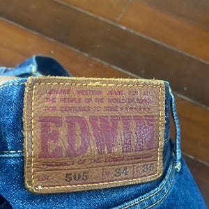 May include: Close-up of a pair of blue denim jeans with a brown leather patch. The patch features the brand name "EDWIN" in red lettering, along with the text "GENUINE WESTERN JEANS FOR ALL THE PEOPLE OF THE WORLD." The jeans have a waist size of 34 inches and a length of 35 inches.