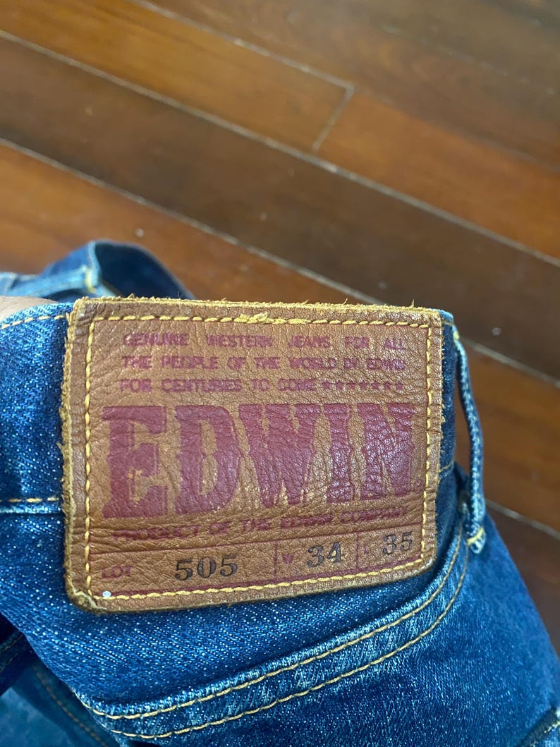 May include: Close-up of a pair of blue denim jeans with a brown leather patch. The patch features the brand name "EDWIN" in red lettering, along with the text "GENUINE WESTERN JEANS FOR ALL THE PEOPLE OF THE WORLD." The jeans have a waist size of 34 inches and a length of 35 inches.