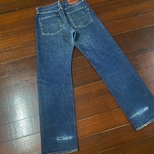 May include: Blue denim jeans with a classic five-pocket design. The jeans feature a faded wash and distressed details at the knees. The Edwin brand label is visible on the back pocket.