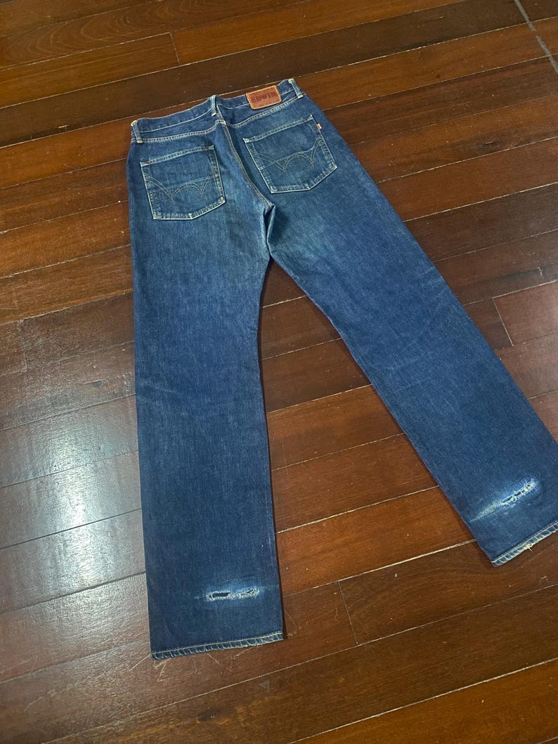 May include: Blue denim jeans with a classic five-pocket design. The jeans feature a faded wash and distressed details at the knees. The Edwin brand label is visible on the back pocket.