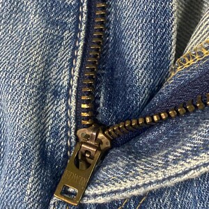 May include: Close-up shot of blue denim jeans, focusing on the metal zipper. The zipper pull features the brand name "EDWIN". The denim fabric has a classic blue wash and visible stitching details.