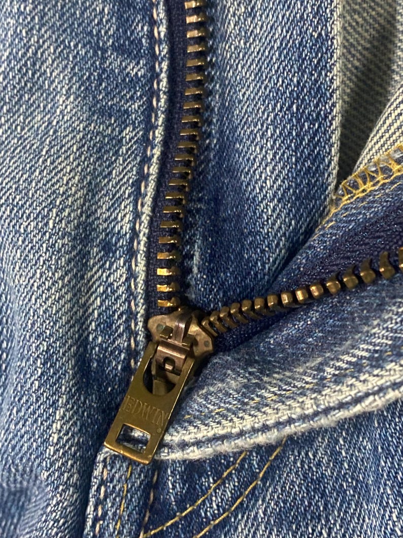 May include: Close-up shot of blue denim jeans, focusing on the metal zipper. The zipper pull features the brand name "EDWIN". The denim fabric has a classic blue wash and visible stitching details.