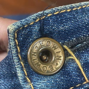 May include: Close-up of a brass button on blue denim fabric. The button is embossed with the word "EDWIN" in a circular pattern. The denim has yellow stitching and a worn appearance.