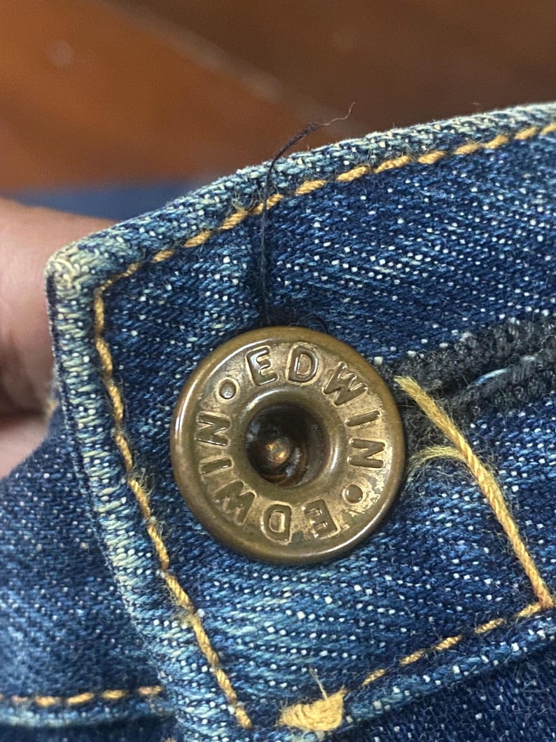 May include: Close-up of a brass button on blue denim fabric. The button is embossed with the word "EDWIN" in a circular pattern. The denim has yellow stitching and a worn appearance.
