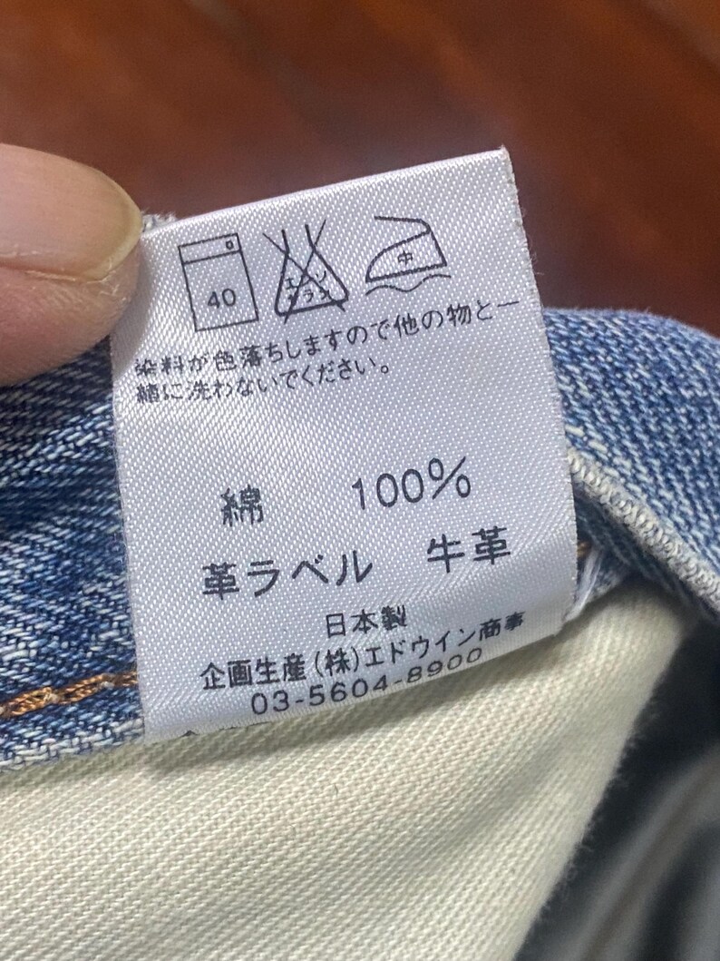 May include: A close-up of a white clothing label with Japanese text and care symbols. The label states the material is 100% cotton and has a leather label. A phone number is also visible.