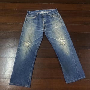 May include: Blue denim jeans with distressed details, including rips on the knees and faded areas. The jeans have a button fly and classic five-pocket design. The denim has a worn appearance.