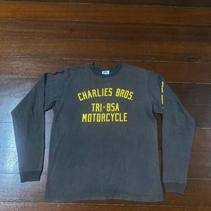 May include: A long-sleeved, dark brown t-shirt with the text "CHARLIES BROS TRI-BSA MOTORCYCLE" in yellow. The shirt has a crew neck and a slightly faded appearance, suggesting a vintage style.
