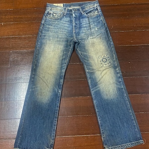May include: Blue denim jeans with a faded wash and a button fly. The jeans have a patch detail on the right leg and a classic five-pocket design. The jeans are laid flat on a wooden surface.