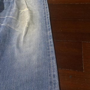 May include: Faded blue denim jeans with a distressed area near the knee. The jeans have a classic five-pocket design and a straight leg cut. The fabric shows signs of wear and tear, giving them a vintage appearance.
