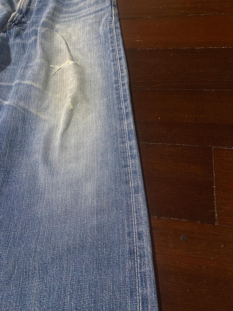 May include: Faded blue denim jeans with a distressed area near the knee. The jeans have a classic five-pocket design and a straight leg cut. The fabric shows signs of wear and tear, giving them a vintage appearance.