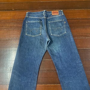 May include: Blue denim jeans with two back pockets and a brown leather patch with the brand name "EDWIN". The jeans are laid flat on a wooden surface, showcasing the back view of the garment.