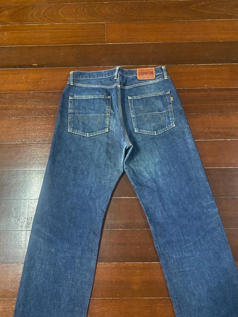 May include: Blue denim jeans with two back pockets and a brown leather patch with the brand name "EDWIN". The jeans are laid flat on a wooden surface, showcasing the back view of the garment.
