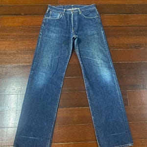 May include: A pair of blue denim jeans with a classic five-pocket design. The jeans have a button fly and a straight leg cut. The denim has a faded appearance, with lighter areas on the thighs and knees. The jeans are laid flat on a wooden surface.