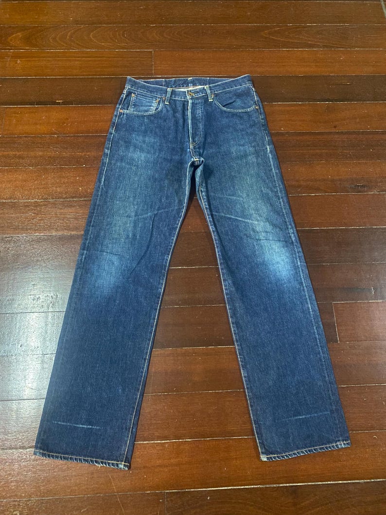 May include: A pair of blue denim jeans with a classic five-pocket design. The jeans have a button fly and a straight leg cut. The denim has a faded appearance, with lighter areas on the thighs and knees. The jeans are laid flat on a wooden surface.