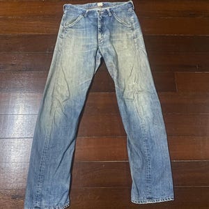 May include: Faded blue denim jeans with a classic five-pocket design. The jeans show signs of wear and tear, with a light wash and subtle distressing. The pants are laid flat on a wooden surface.