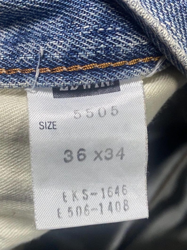 May include: Close-up of a white label from blue denim jeans. The label shows the size as 36 x 34 inches (91.4 x 86.4 cm), along with the number 5505 and other codes. The jeans are made of denim.