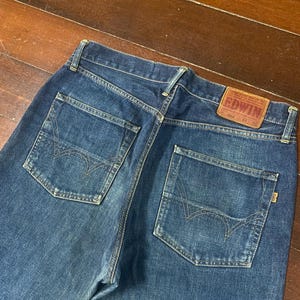 May include: Dark blue denim jeans with two back pockets and a leather patch. The jeans have a classic five-pocket design and a straight leg fit. The Edwin brand label is visible on the back.