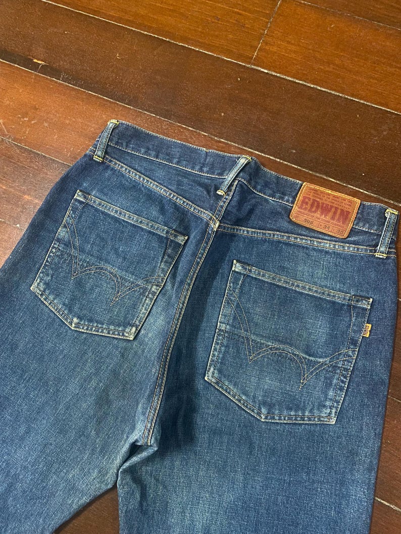 May include: Dark blue denim jeans with two back pockets and a leather patch. The jeans have a classic five-pocket design and a straight leg fit. The Edwin brand label is visible on the back.