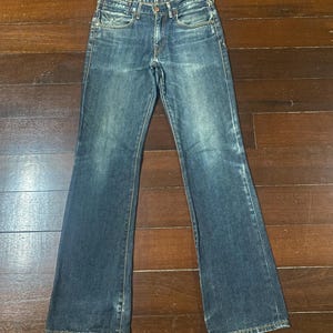 May include: Dark wash denim jeans with a classic five-pocket design and a slight flare at the hem. The jeans have a button and zipper closure and are laid flat on a wooden surface.