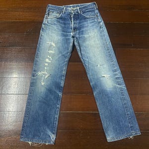 May include: Faded blue denim jeans with distressed details, including rips and wear marks. The jeans have a button fly and classic five-pocket styling. The denim has a vintage wash and a slightly worn appearance.