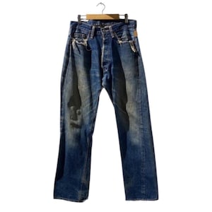 Op de afbeelding: Verbleekte blauwe spijkerbroek met distressed details, waaronder scheuren en versleten plekken. De jeans worden getoond op een houten hanger, wat een vintage of versleten esthetiek laat zien. De jeans hebben een knoopsluiting en een klassiek vijf-zakken-ontwerp.