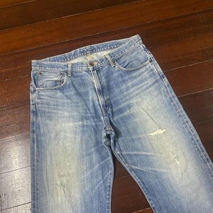 May include: Faded blue denim jeans with a classic five-pocket design. The jeans show signs of wear, including light distressing and fading, giving them a vintage look. The jeans are laid flat on a wooden surface.