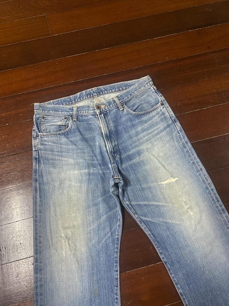 May include: Faded blue denim jeans with a classic five-pocket design. The jeans show signs of wear, including light distressing and fading, giving them a vintage look. The jeans are laid flat on a wooden surface.