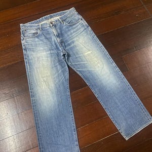 May include: Light blue denim jeans with a classic five-pocket design. The jeans have a straight leg cut and show signs of wear, including light fading and a small distressed area on the front. The jeans are laid flat on a wooden surface.
