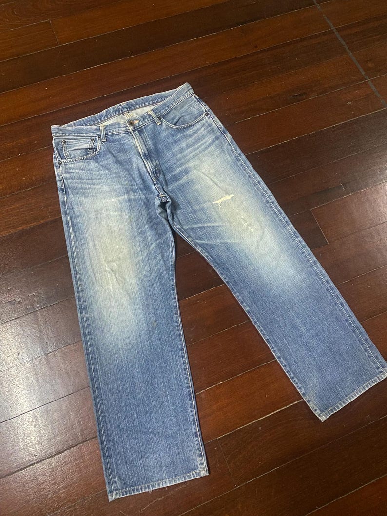 May include: Light blue denim jeans with a classic five-pocket design. The jeans have a straight leg cut and show signs of wear, including light fading and a small distressed area on the front. The jeans are laid flat on a wooden surface.