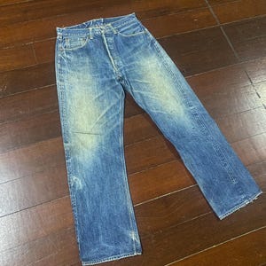 May include: Faded blue denim jeans with a button fly. The jeans have a classic five-pocket design and a slightly worn appearance. The denim has a vintage wash with lighter areas on the thighs and knees. The jeans are laid flat on a wooden surface.