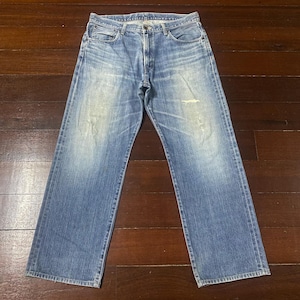 May include: Light blue denim jeans with a faded wash and a relaxed fit. The jeans feature a classic five-pocket design, a button and zip closure, and a distressed area on the right leg. The jeans are laid flat on a wooden surface.