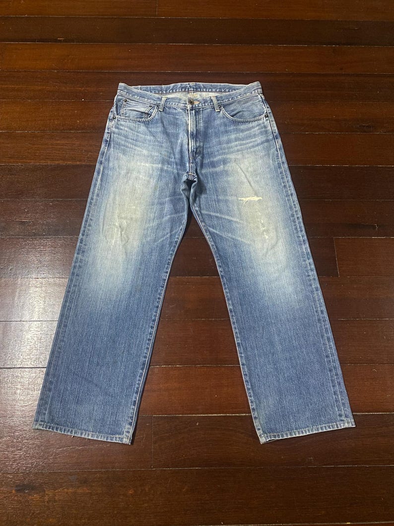 May include: Light blue denim jeans with a faded wash and a relaxed fit. The jeans feature a classic five-pocket design, a button and zip closure, and a distressed area on the right leg. The jeans are laid flat on a wooden surface.