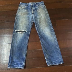 May include: Faded blue denim jeans with a distressed look. The jeans have a button fly, classic five-pocket design, and a large rip on the left knee. The denim shows signs of wear and fading, giving them a vintage aesthetic.