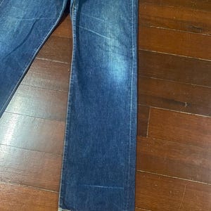 May include: Close-up of blue denim jeans. The jeans have a classic five-pocket design and a straight leg cut. The denim has a faded appearance, with lighter areas on the thighs and knees. The jeans are laid flat on a wooden surface.