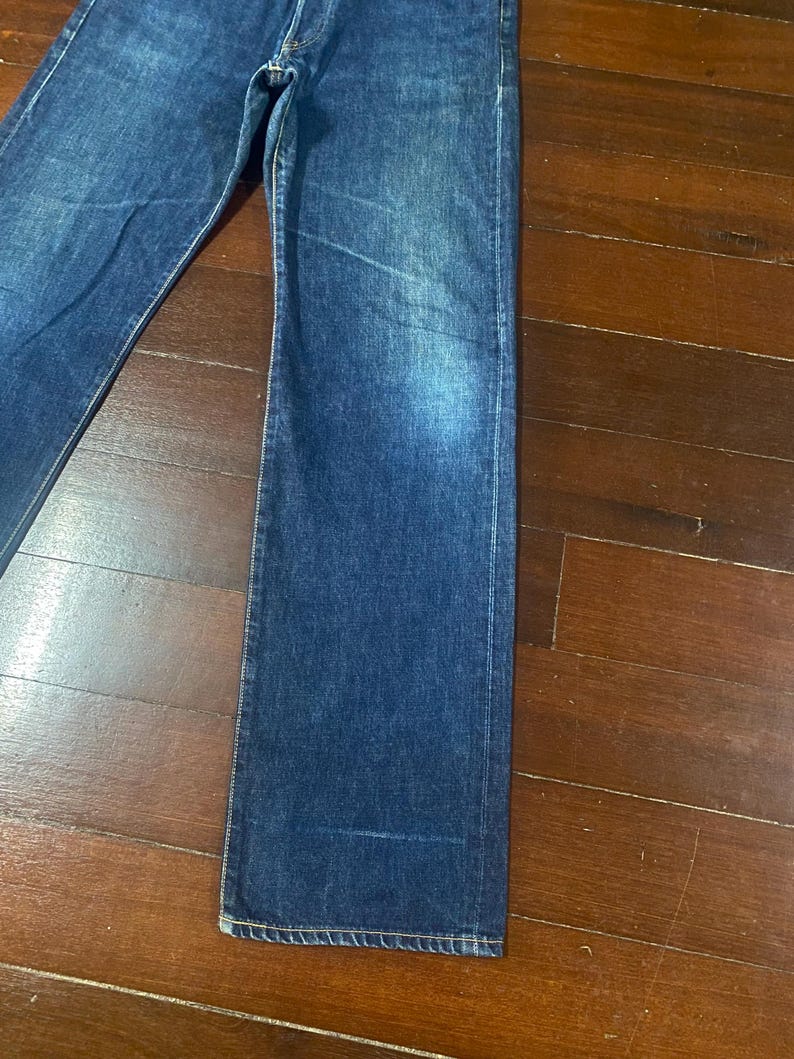 May include: Close-up of blue denim jeans. The jeans have a classic five-pocket design and a straight leg cut. The denim has a faded appearance, with lighter areas on the thighs and knees. The jeans are laid flat on a wooden surface.
